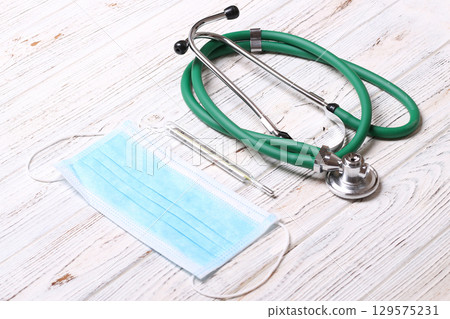 Top view of stethoscope, protective mask and mercury thermometer on white wooden background. Medical instruments concept 129575231