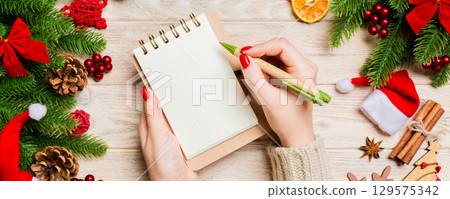 Top view of female hand writing in a notebook on wooden Christmas background. Fir tree and festive decorations. Wish list. New Year concept 129575342