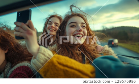 Two friends enjoying a drive on a road trip. One captures the moment with a phone while the other laughs joyfully. Freedom and friendship captured in this perfect shot. Generative AI 129576986