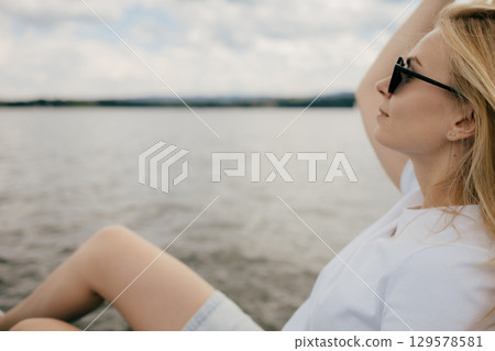 Blond woman wearing sunglasses sits on boat, soaking up summer sun while enjoying serene lake and picturesque hills in background, embodying relaxation and tranquility. Blond woman relaxing on boat 129578581