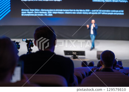 Conference audience watching speaker on stage with cameras recording the presentation 129579810