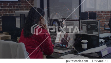 Attending virtual meeting Indian professional in red blazer using laptop at office, with smartphone 129580152