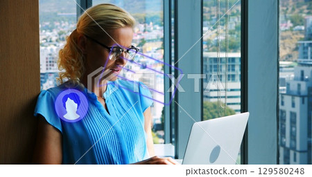 woman in blouse and glasses typing on laptop near office window, with holographic icons, copy space 129580248