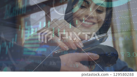 Image of financial data and graphs over happy caucasian woman using payment terminal 129580288