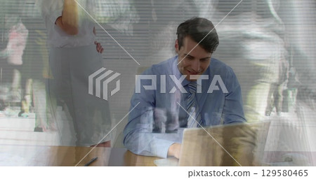 Man in shirt and tie typing on laptop at office desk, with window blinds showing pedestrians 129580465