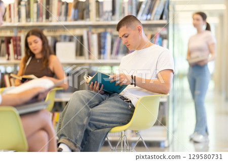Male student sitting with book on chair in library 129580731