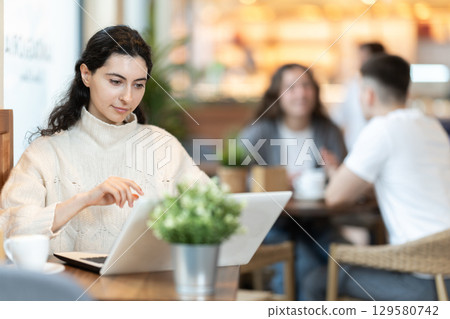 In cozy cafe hall, freelance girl works with laptop and drinks coffee. In cozy cafe hall, freelance girl works with laptop and drinks coffee. 129580742