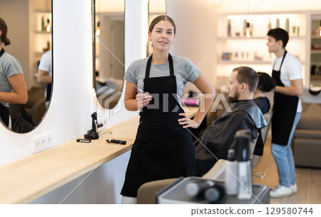 Girl hairdresser stylist stands near workplace with tool in hands. Girl hairdresser stylist stands near workplace with tool in hands. 129580744