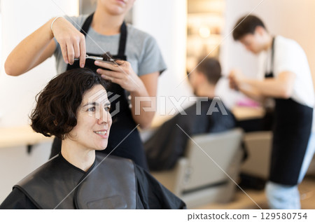 Young woman barbershop employee stands next to female client, discussing details of haircut 129580754