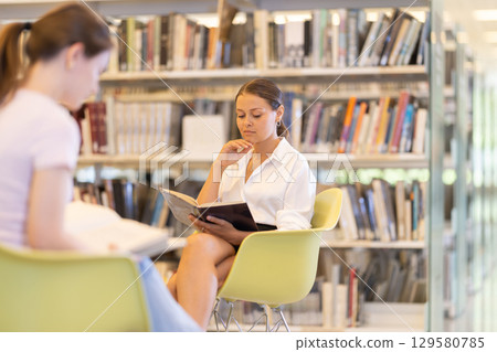 Female student reading book on chair in library Female student reading book on chair in library 129580785