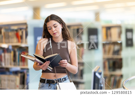 Young female student reading book in library Young female student reading book in library 129580815