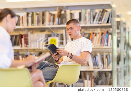 Male student reading book on chair in library Male student reading book on chair in library 129580818