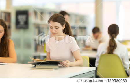 Female student sits at table in library and studies, reads books and takes notes 129580983