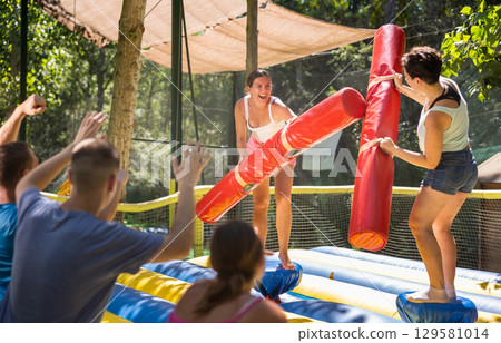 Young women having funny wrestling by inflatable logs in outdoor amusement playground 129581014