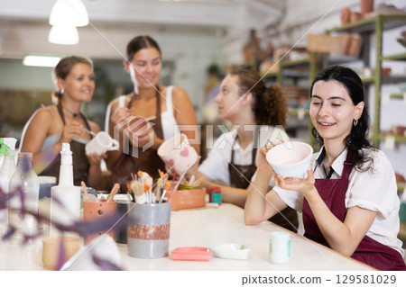 Young woman sits near table and decorates clay ceramic craft vase, plate 129581029