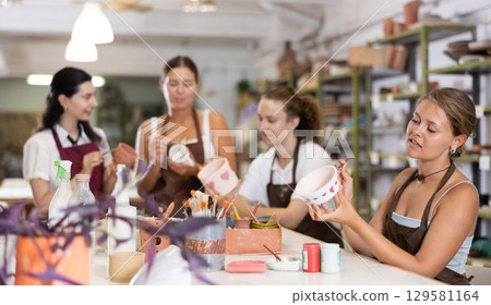 Young woman sits near table and decorates clay ceramic craft vase, plate Young woman sits near table and decorates clay ceramic craft vase, plate 129581164