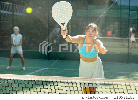 Happy woman learning to play padel game on tennis court outdoor. Other athletes are training in background Happy woman learning to play padel game on tennis court outdoor. Other athletes are training in background 129581192