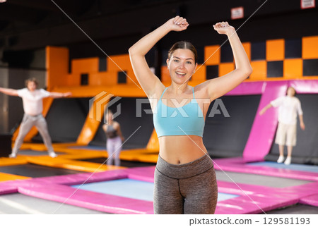 Happy young European female in blue top and gray leggings posing on trampolines during active weekend free time in sports center 129581193
