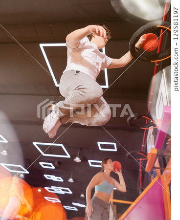 Encouraged young guy in active wear throwing ball in basketball basket while jumping on trampolines in entertainment center 129581197