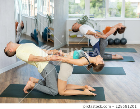 Lovers of active lifestyle perform pair yoga supported backbend on knee in gym 129581201