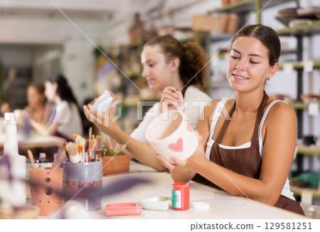 Young woman sits near table and decorates clay ceramic craft vase, plate 129581251