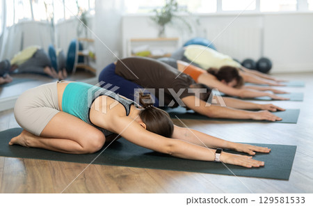 Lovers of active lifestyle on yoga mat perform doing version of child pose in gym Lovers of active lifestyle on yoga mat perform doing version of child pose in gym 129581533