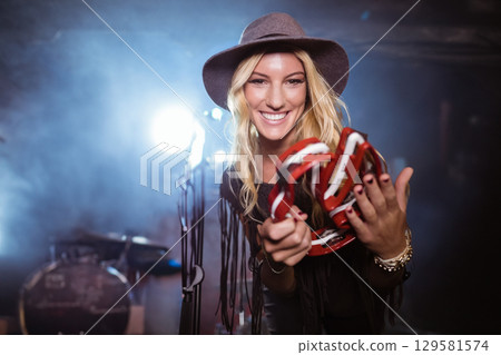Female musician standing on stage holding red-and-white tambourine with stage lights glowing behind Female musician standing on stage holding red-and-white tambourine with stage lights glowing behind 129581574