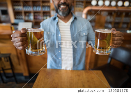 Mature Indian man wearing denim shirt holding two frothy beer mugs at wooden bar counter 129581578
