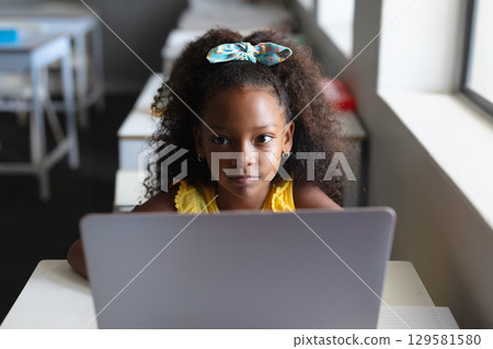 African American schoolgirl sitting at school desk with headband working on laptop near window 129581580