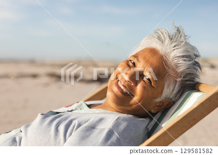 Senior woman reclining on wooden lounge chair with striped cushion on beach in sun, copy space Senior woman reclining on wooden lounge chair with striped cushion on beach in sun, copy space 129581582