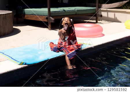 African American girl sitting wrapped in red towel at pool edge licking popsicle under sunlight African American girl sitting wrapped in red towel at pool edge licking popsicle under sunlight 129581592