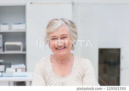 Smiling senior female sitting at desk in home office sorting paperwork near printer and shelving 129581593