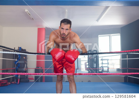 Male boxer leaning forward against boxing ring rope in gym wearing red gloves and shorts Male boxer leaning forward against boxing ring rope in gym wearing red gloves and shorts 129581604