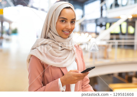Woman wearing beige headscarf and pink blazer holding smartphone and smiling in mall atrium 129581608