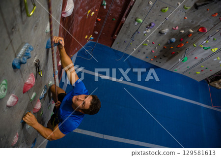 Male climber wearing blue shirt ascending gym wall with multicolored holds, safety rope, copy space 129581613