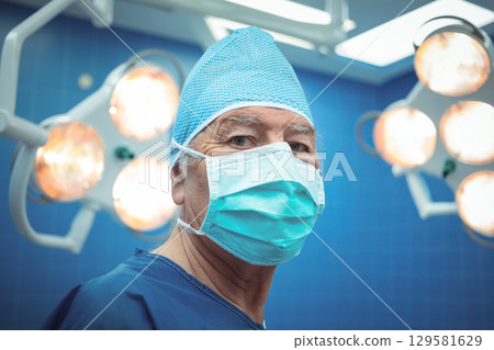 Senior male surgeon standing in operating room under overhead surgical lights wearing mask and cap Senior male surgeon standing in operating room under overhead surgical lights wearing mask and cap 129581629