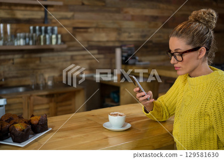 Woman wearing chunky sweater scrolling smartphone at cafe bar with latte, muffins copy space Woman wearing chunky sweater scrolling smartphone at cafe bar with latte, muffins copy space 129581630