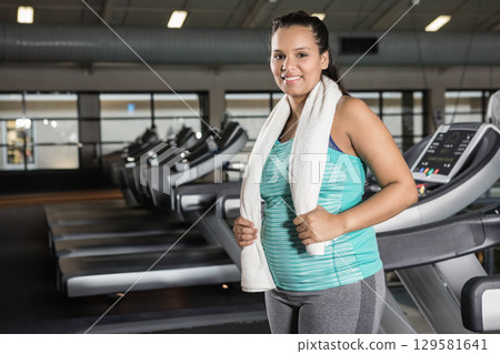 Woman standing beside treadmill wearing turquoise top, leggings with white towel in gym, copy space 129581641