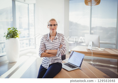 Mature woman working on open laptop at modern office near planter and monitor in business attire 129581648
