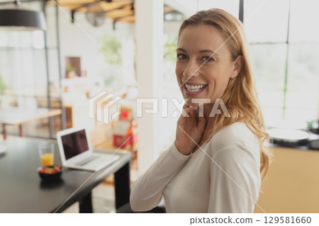 Woman standing beside dining table under pendant lamp in open-plan home with laptop, juice, fruit 129581660