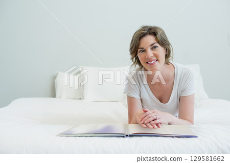 Mid adult woman lying on white linens with pillows in bedroom reading book and wearing t-shirt 129581662