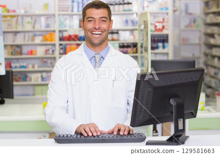 Male pharmacist standing at pharmacy counter typing on keyboard with shelves of medication Male pharmacist standing at pharmacy counter typing on keyboard with shelves of medication 129581663