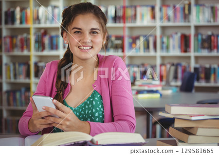 Teenage girl wearing pink cardigan sitting at library table holding smartphone and open book 129581665