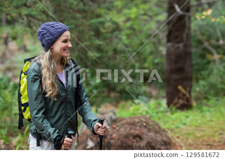 Woman gripping trekking poles and hiking along forest trail in waterproof jacket, copy space 129581672