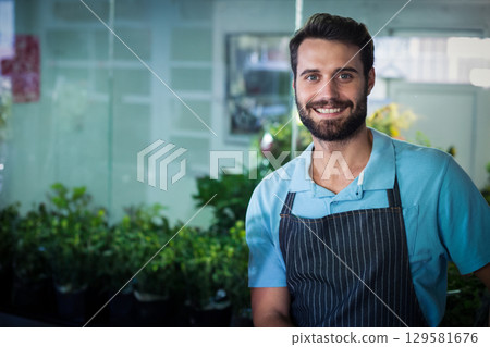 Man wearing striped apron in plant shop smiling at camera among potted plants, copy space 129581676