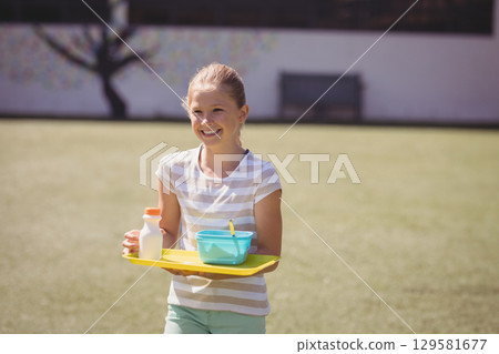 Female child holding yellow plastic tray with bottle, turquoise bowl and spoon on grassy lawn 129581677
