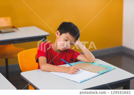 Asian boy resting head on hand writing in colorful workbook at school desk with pencil 129581681