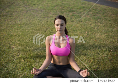 Woman sitting cross-legged on park lawn practicing meditation wearing pink top with fitness tracker Woman sitting cross-legged on park lawn practicing meditation wearing pink top with fitness tracker 129581682