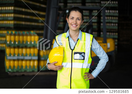 Female warehouse supervisor standing in storage wearing safety vest holding hardhat with forklift 129581685