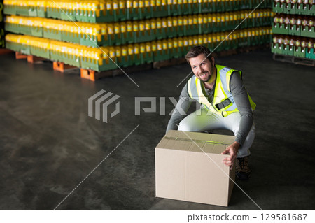 Male warehouse worker wearing vest kneeling in warehouse lifting box beside crates, copy space 129581687